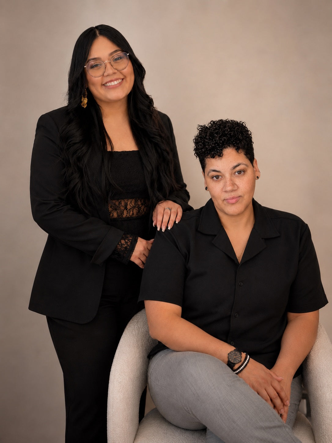 Two women in black outfits posing together in front of a neutral beige studio backdrop. One woman stands on the left wearing a black blazer over a black top, and the other sits on a light gray chair on the right wearing a black short-sleeve collared shirt and light gray pants.